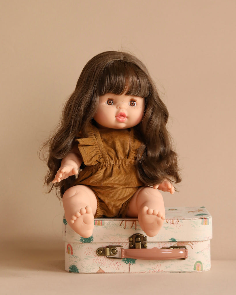 A Minikane Baby Doll (13") - Valentine, featuring long brown hair and exuding a natural vanilla scent, is seated on a patterned suitcase. The doll is dressed in a brown dress against a neutral beige background.