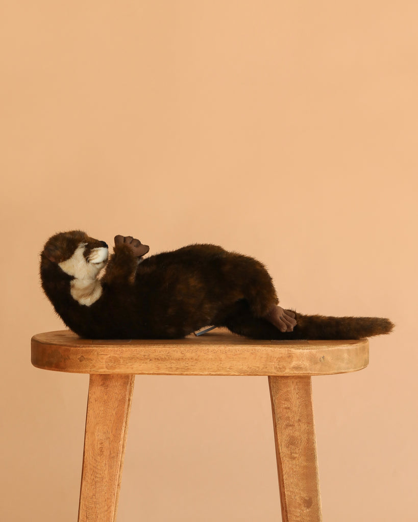 The Otter Mama Stuffed Animal lies on its back on a wooden stool against a plain beige background.