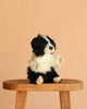 The Border Collie Dog Puppet, a hand-sewn black and white plush with pink paws, sits upright on a wooden stool against a plain beige background.