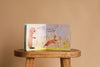 The interactive board book "I Love You So" by Patricia Hegarty is open on a wooden stool, showing two illustrated baby rabbits in a field with the text, "Mummy rabbit watches her baby goes skippety-hop." The background is plain beige.