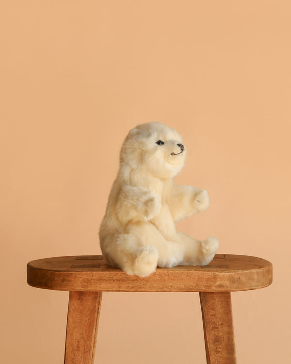 A fluffy white Polar Bear Puppet sits upright on a wooden stool against a plain peach background, showcasing the lifelike charm of realistic plush animals.