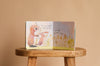 The children’s interactive board book "I Love You So" by Patricia Hegarty sits open on a wooden stool, showing two illustrated baby rabbits in tall grass with the text: “When you scamper back to me—we’ll hug and never stop.”.