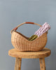 A Basket of Lavender Felt Flowers is placed on a rustic wooden stool against a plain gray background.