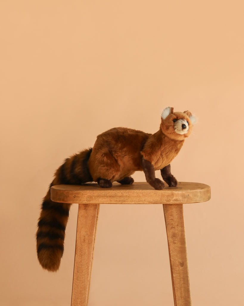 A hand-sewn plush mongoose stuffed animal with realistic features and a bushy striped tail stands on a wooden stool against a plain beige background.