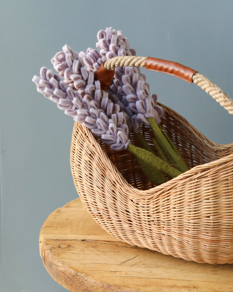 A Basket of Lavender Felt Flowers rests on a wooden surface. The half moon basket contains several stems of handmade felt flowers in purple, accented with green felt leaves. The background is a soft, muted blue wall.