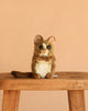 The Tarsier Baby Stuffed Animal, a hand-sewn plush with brown and white fur, big ears, round eyes, and a long tail, sits on a wooden bench against a plain beige background.