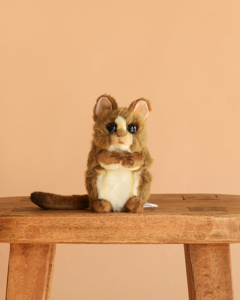 The Tarsier Baby Stuffed Animal, a hand-sewn plush with brown and white fur, big ears, round eyes, and a long tail, sits on a wooden bench against a plain beige background.
