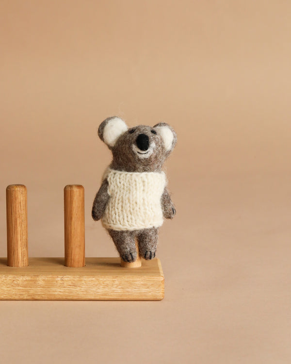 The Felt Finger Puppet: Baby Koala, dressed in a white knitted sweater, stands next to a wooden holder with two vertical pegs against a plain beige background.