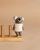 The Felt Finger Puppet: Baby Koala, dressed in a white knitted sweater, stands next to a wooden holder with two vertical pegs against a plain beige background.