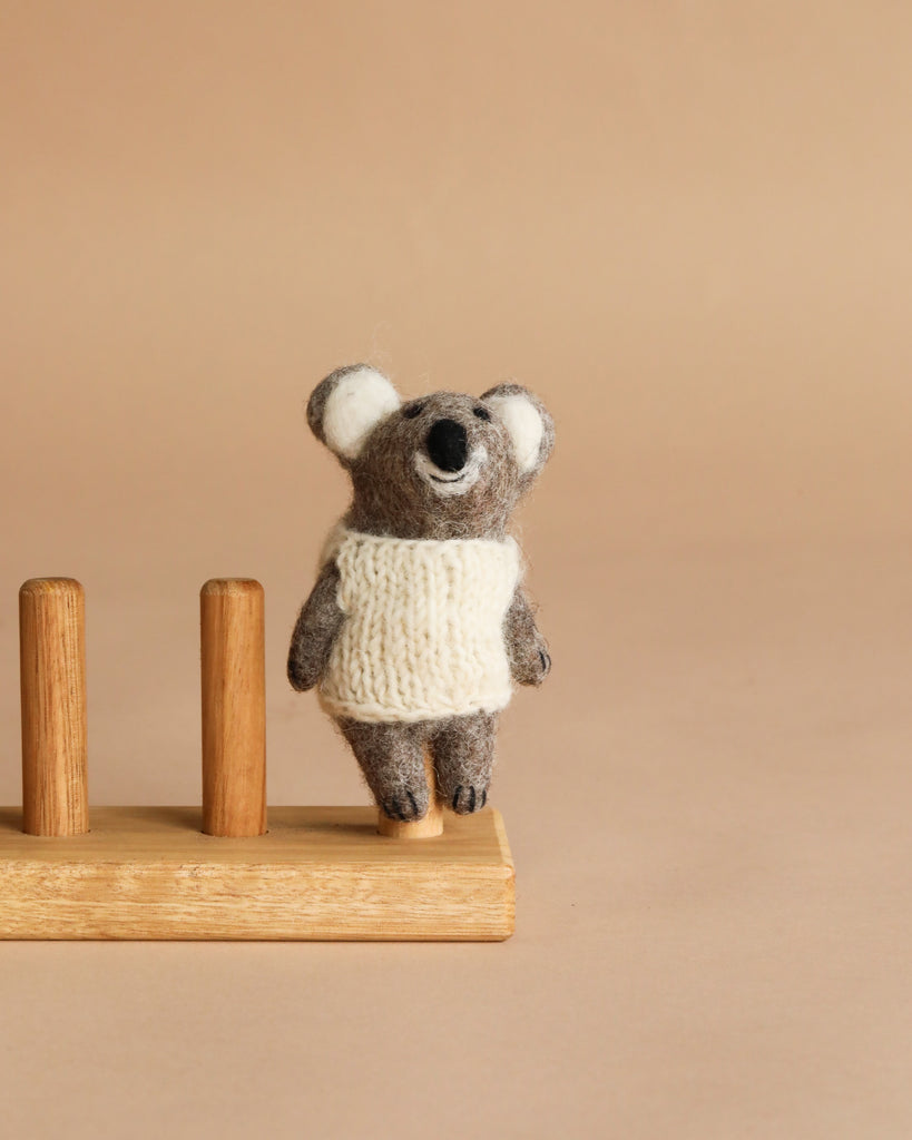 The Felt Finger Puppet: Baby Koala, dressed in a white knitted sweater, stands next to a wooden holder with two vertical pegs against a plain beige background.