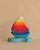 Grimm's Stacking Tower Sailboat features semi-circular wooden blocks, arranged by size and color in a rainbow teardrop shape on a light blue base—perfect for fine motor skill development against a beige background.