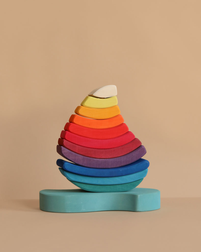Grimm's Stacking Tower Sailboat features semi-circular wooden blocks, arranged by size and color in a rainbow teardrop shape on a light blue base—perfect for fine motor skill development against a beige background.