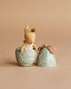 The Maileg Easter Egg with Micro Rabbit, Small - Dusty Blue features a soft rabbit in a polka-dot outfit inside a blue floral egg-shaped container, placed beside its ribbon-tied lid on a beige background.