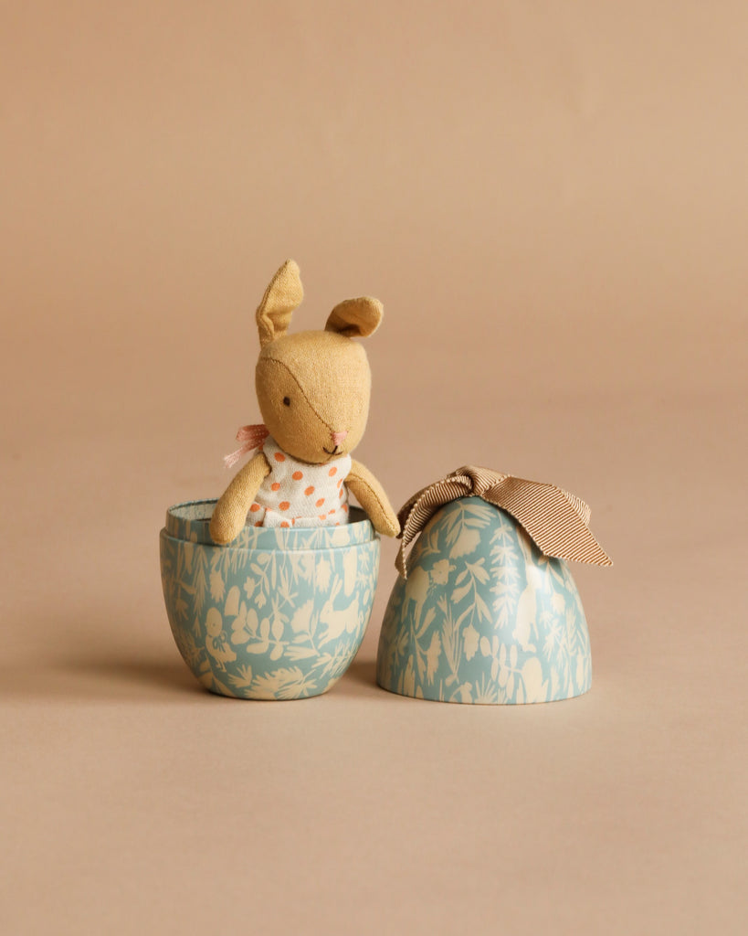 The Maileg Easter Egg with Micro Rabbit, Small - Dusty Blue features a soft rabbit in a polka-dot outfit inside a blue floral egg-shaped container, placed beside its ribbon-tied lid on a beige background.
