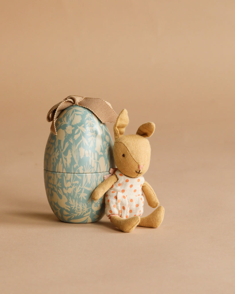The Maileg Little Rabbit’s Rocking Chair Set features a small rabbit in a polka dot dress next to a floral-patterned Maileg Easter Egg with a ribbon, all displayed on a beige background.