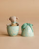 The Maileg Easter Egg & Friends Set features a small stuffed Micro Lamb with a green ribbon nestled inside a blue-green floral-patterned egg container, its lid placed nearby on a beige background.