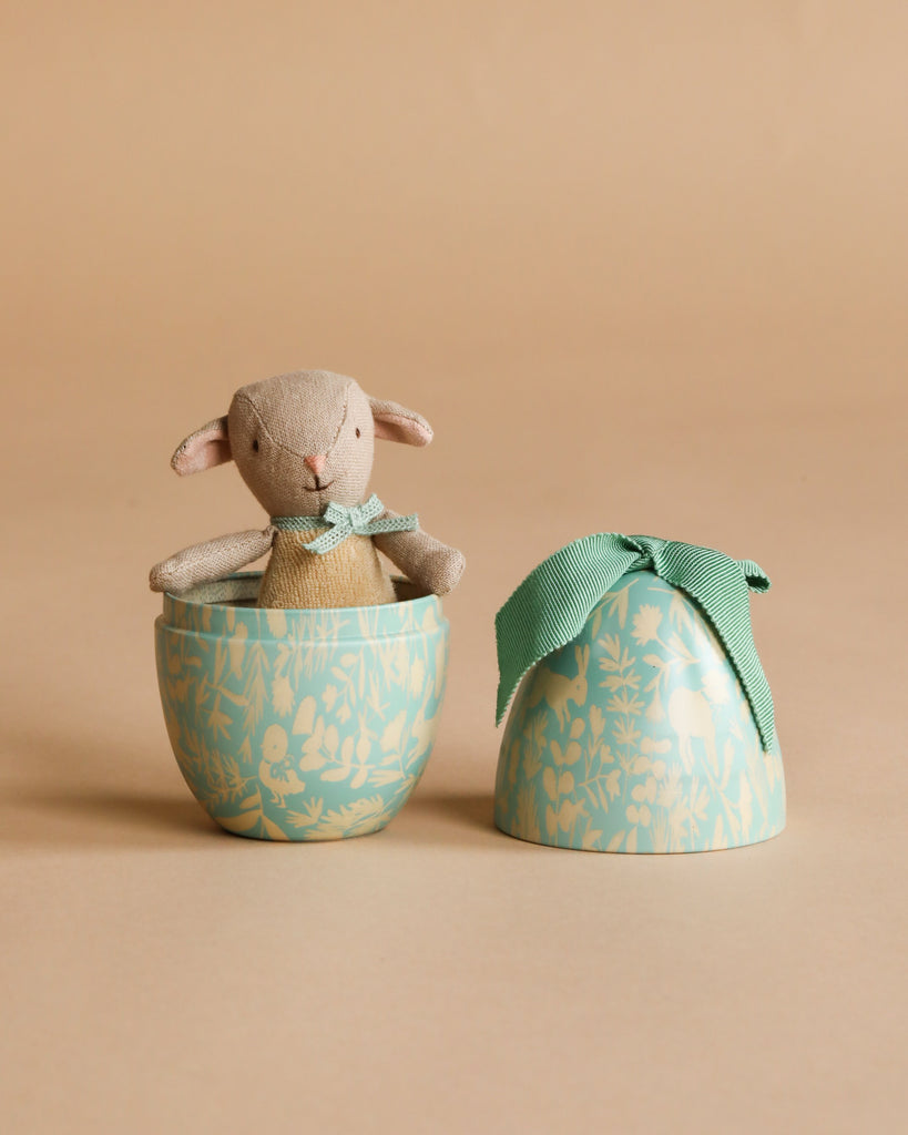 The Easter Basket Gift Set features a small stuffed lamb with a green ribbon, sitting inside a decorative egg-shaped container with the lid beside it on a beige background.