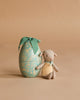 The Maileg Easter Egg with Micro Lamb, Small - Dusty Mint features a soft lamb with a green bow and yellow apron beside a light blue floral egg with a green ribbon, all set against a plain beige background.