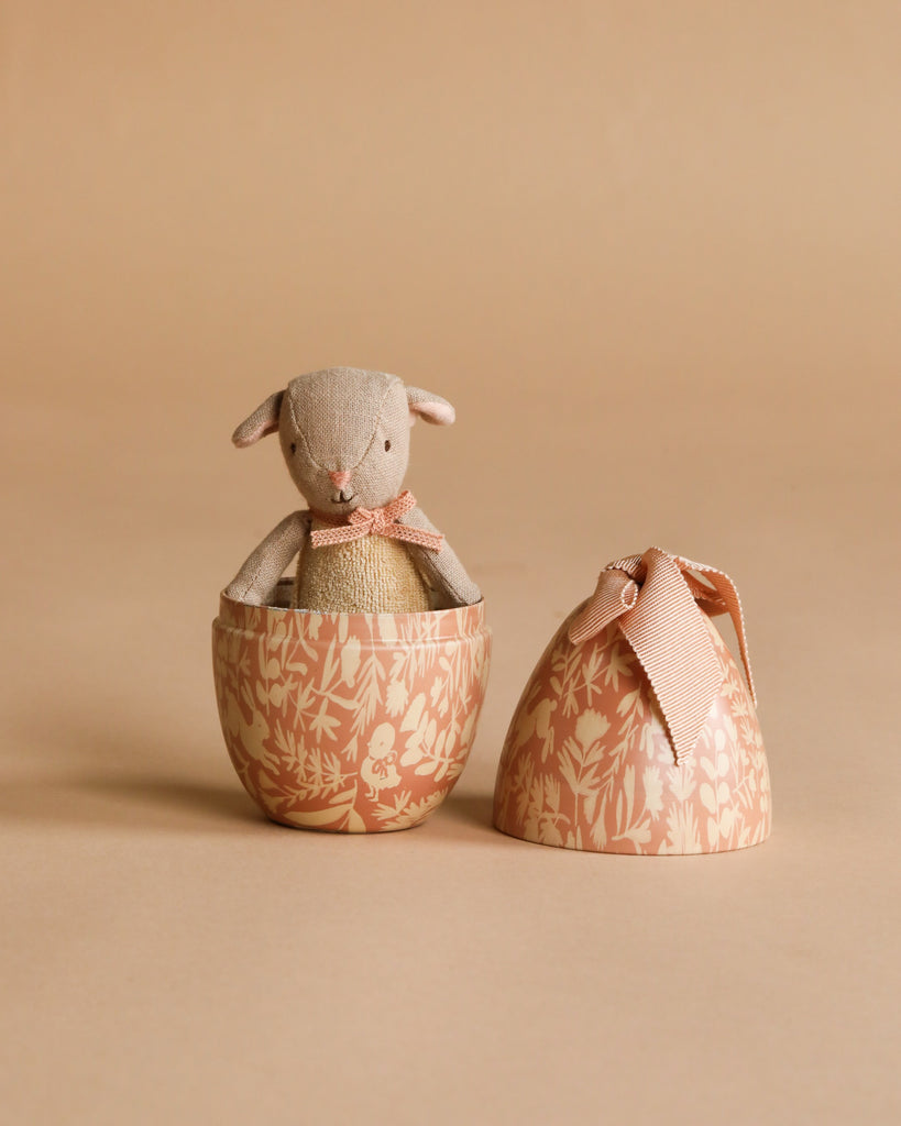 A Maileg Little Lambs Kitchen Set featuring a plush lamb with a pink ribbon is displayed inside a floral Maileg Easter Egg, with the matching lid nearby, all set against a beige background.