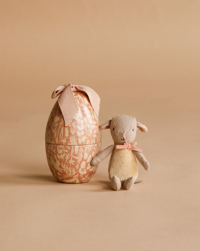 A small plush mouse with a pink ribbon sits beside the Maileg Easter Egg with Lamb & Extra Clothing—an egg-shaped box with floral patterns and a fabric bow—both set against a neutral beige background.