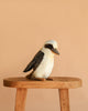The Australian Kookaburra Bird Stuffed Animal, featuring brown, white, and cream colors, sits on a wooden stool against a plain light beige background—one of HANSA's realistic plush toys.