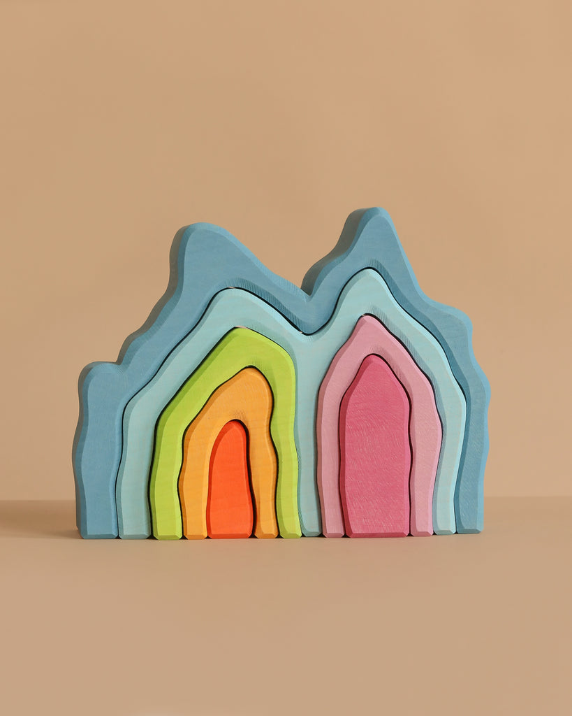 Pastel-colored wooden arches from Grimm's Blue Cave Arch Stacking Toy, resembling abstract mountains, stacked on a plain beige background. The arches come in blue, green, yellow, orange, pink, and purple.