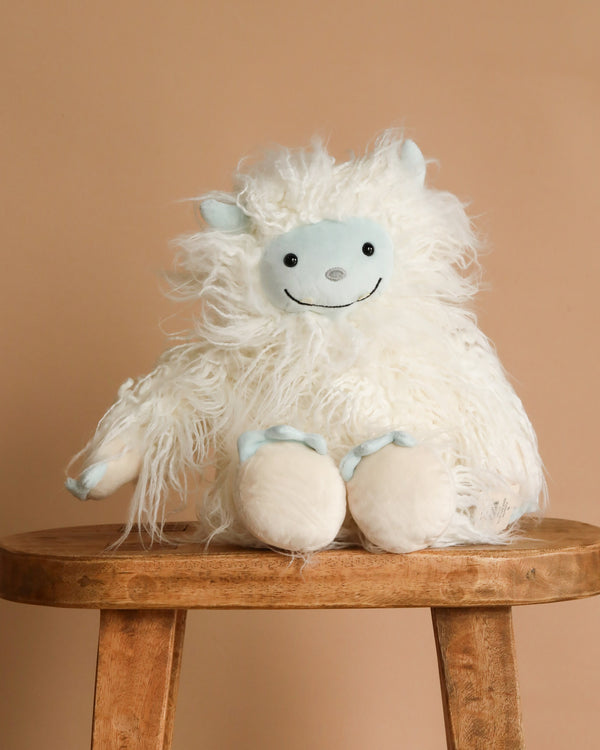 Mon Ami - Yowie the Yeti Plush Toy, featuring a fluffy white body, light blue face, hands, feet, and small ears, sits on a wooden stool with a beige background. This soft plush has a friendly smile and shaggy texture.