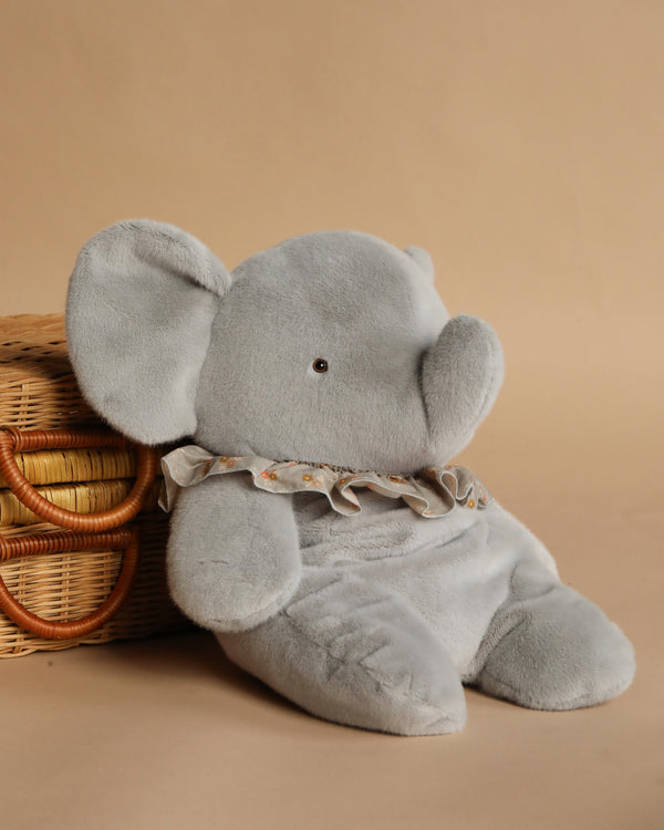 The Maileg Elephant Plush, Big - Blue/Grey, a soft stuffed animal with a ruffled collar, sits upright beside a woven basket on a beige background.