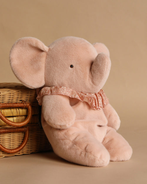 The Maileg Elephant Plush, Big - Powder, wearing a ruffled collar, sits beside a wicker basket against a beige background.