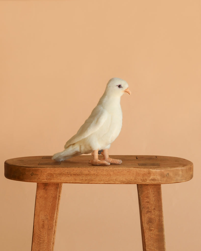 The White Dove Bird Stuffed Animal, featuring a peach beak and legs, stands on a simple wooden stool against a plain beige background.