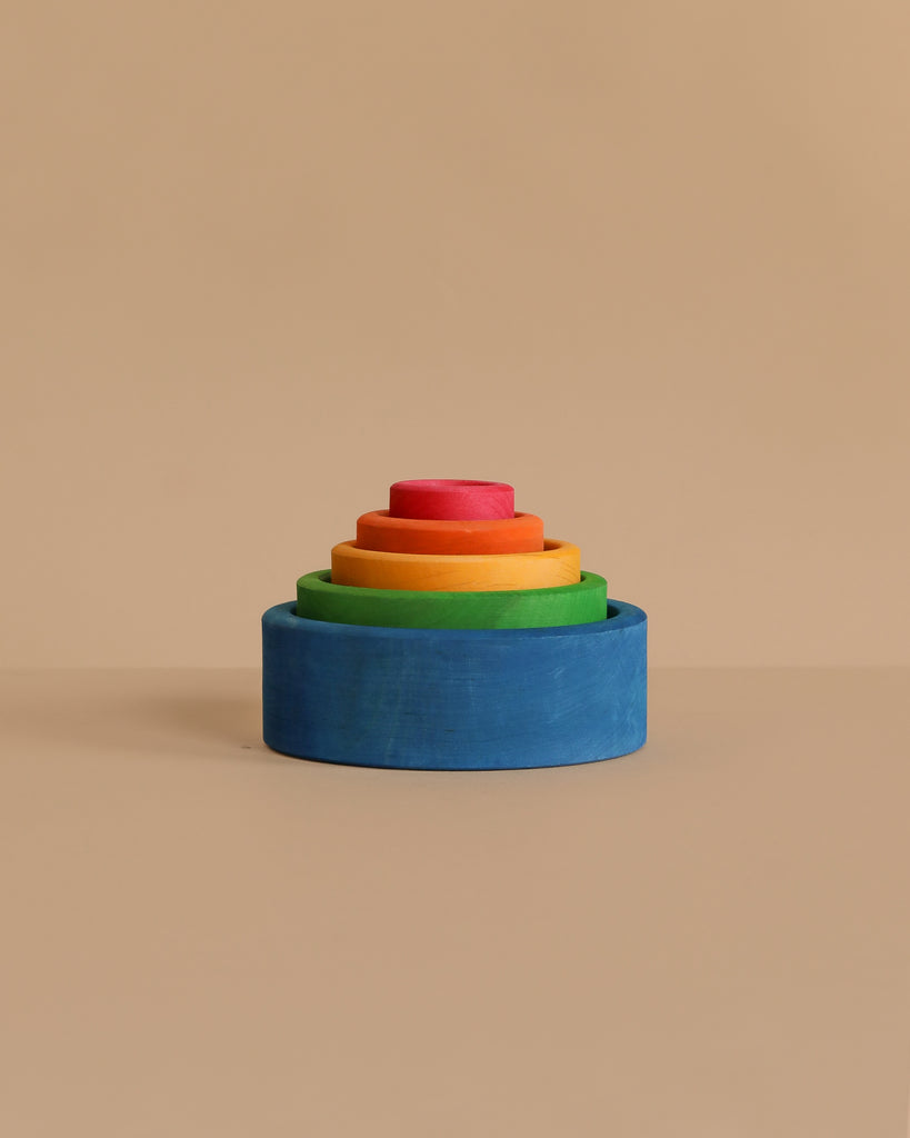 Grimm's Rainbow Stacking Bowls Blue to Red features colorful wooden bowls in graduated sizes, finished with a natural non-toxic stain, and arranged in a vibrant stack against a plain beige background.