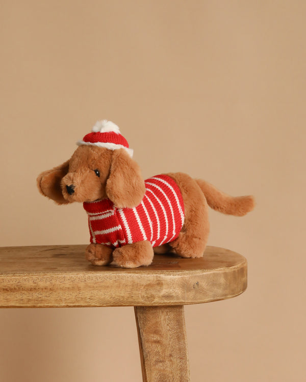 The Mon Ami Jingle Holiday Dachshund Dog Plush Toy, wearing a red and white striped sweater and matching hat, sits on a wooden stool against a plain beige background.