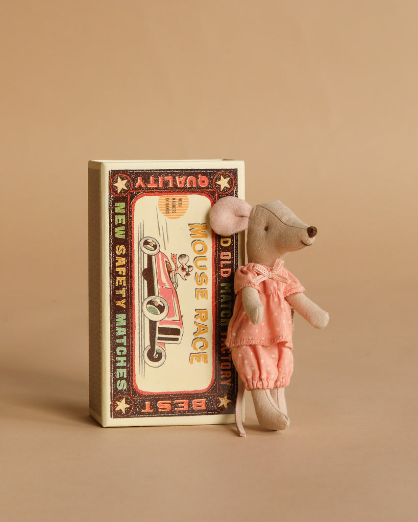 A Maileg Big Sister Bathroom Set features a small mouse in a pink outfit leaning against a vintage-style matchbox with a mouse and car illustration on the front, set against a plain beige background.