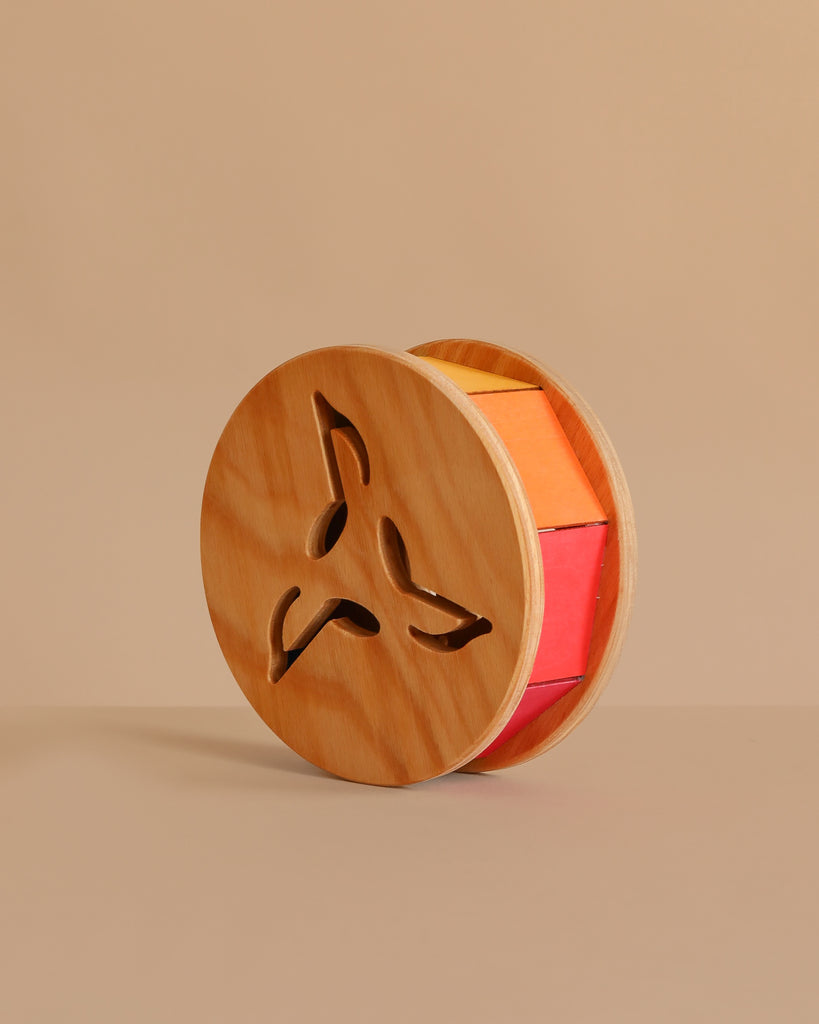 Grimm's Sound and Color Wheel is a wooden baby toy with circular sides featuring geometric cutouts and colorful blocks inside, ideal for motor skills development. Shown here against a plain beige background.