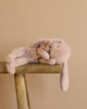 A Maileg Sleeping Bunny in Carry Cot, soft and light pink, rests on its side on a wooden bench against a beige background, adorned with a fabric flower near its neck.