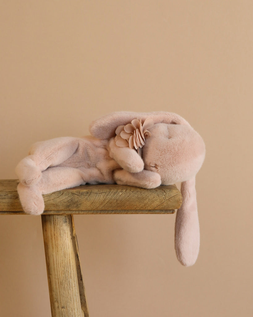 A Maileg Sleeping Bunny in Carry Cot, soft and light pink, rests on its side on a wooden bench against a beige background, adorned with a fabric flower near its neck.