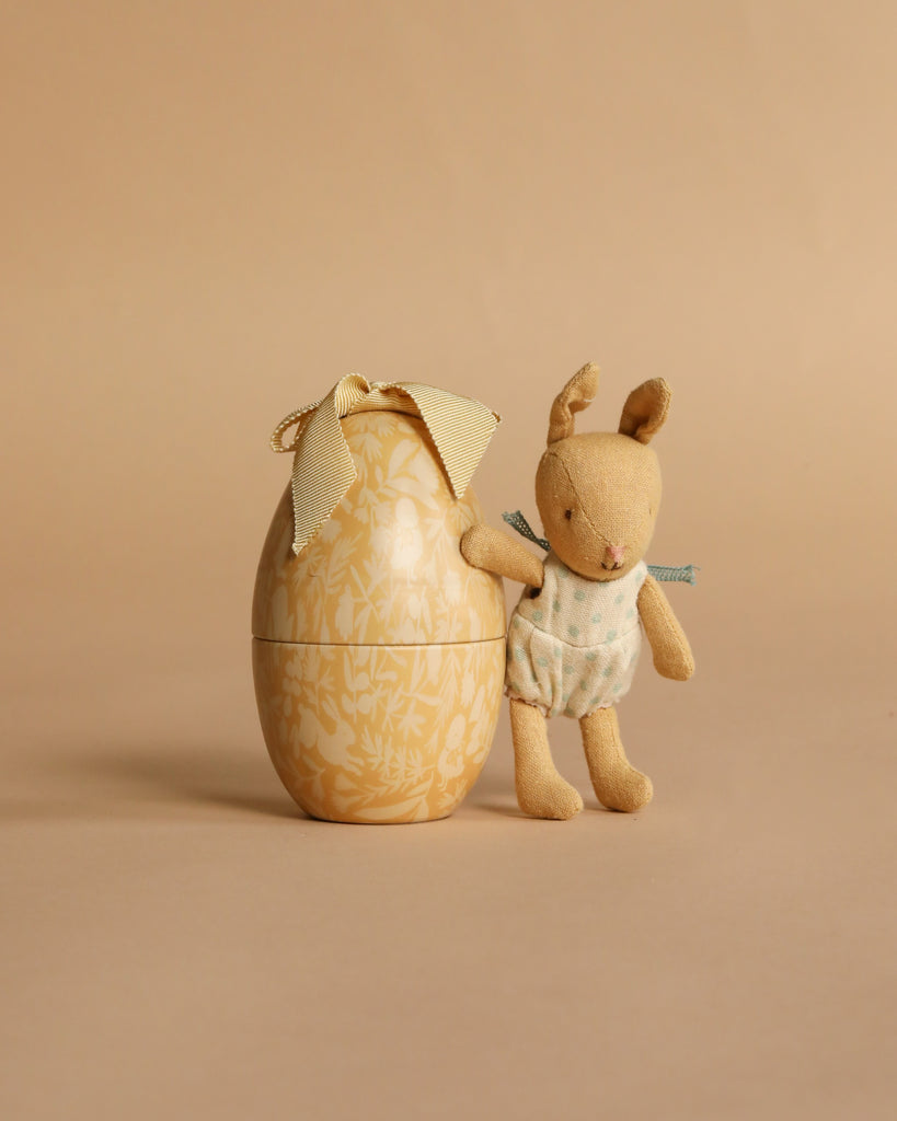 The Maileg Suitcasa Furnished Set (ships in one week) features charming dollhouse accessories, including a small plush bunny in a blue polka-dot outfit and a decorative yellow egg with a floral pattern and ribbon bow on a beige background.
