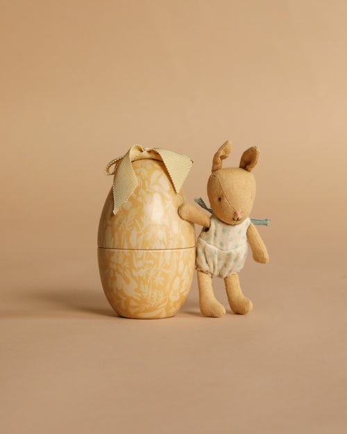 The Maileg Easter Egg with Micro Rabbit, Small - Cream Yellow features a soft rabbit in a polka-dot outfit standing next to a decorative cream yellow egg with a ribbon bow, set against a plain beige background.