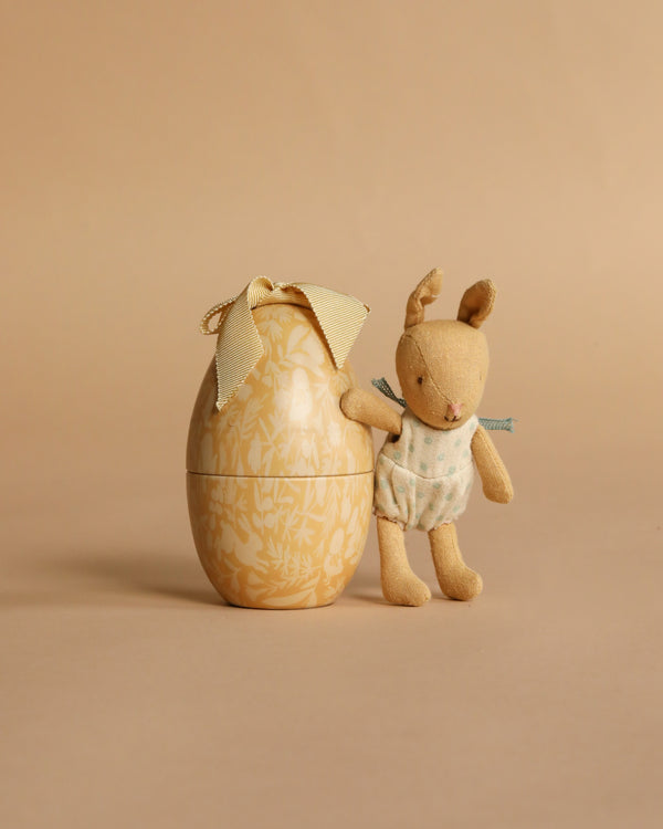 The Maileg Easter Egg with Micro Rabbit, Small - Cream Yellow features a soft rabbit in a polka-dot outfit standing next to a decorative cream yellow egg with a ribbon bow, set against a plain beige background.