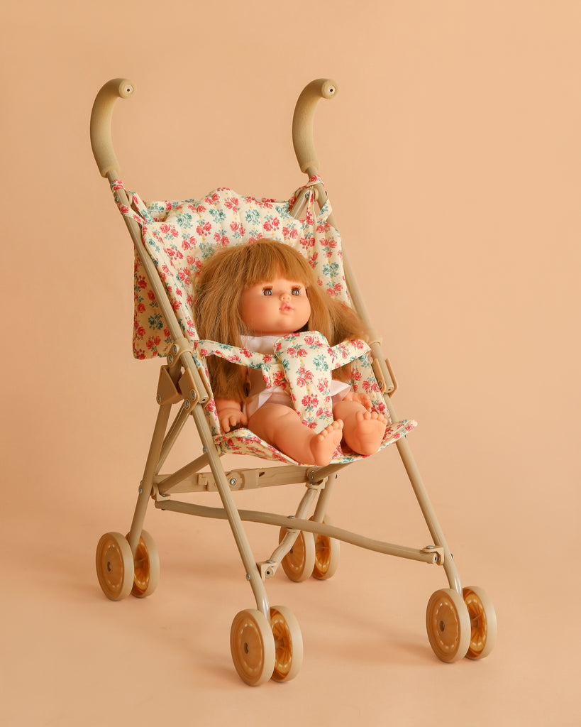 The Minikane Baby Doll (13") – Nina with Clothing and Stroller sits in a beige stroller with a floral print seat and backrest, set against a peach-colored background.