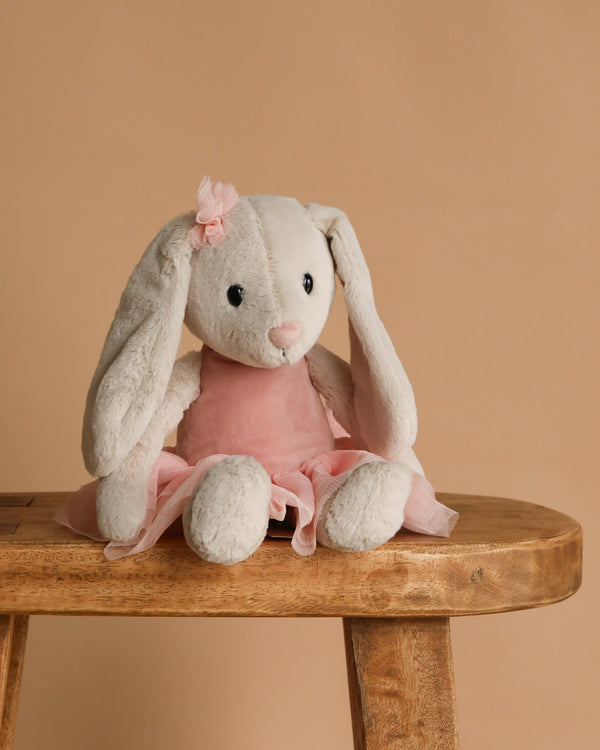 The Steiff Hoppie Rabbit Ballet, dressed in a pink tutu and bow, sits on a wooden bench against a beige background.