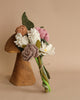 A bouquet of Felt Flower Bouquet - Daydream in various colors including pink, white, and brown, tied with twine and resting on a wooden display stand against a tan background.