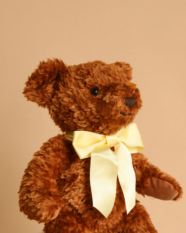 The Steiff Cosy Year Bear 2026 13" is a brown plush teddy with a fluffy texture and a large pale yellow satin bow around its neck, shown against a plain beige background.