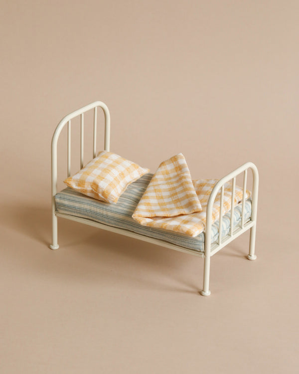A Maileg Miniature Bed with a striped blue mattress and two yellow checkered pillows against a beige background, perfect for Maileg friends' sleep-overs.