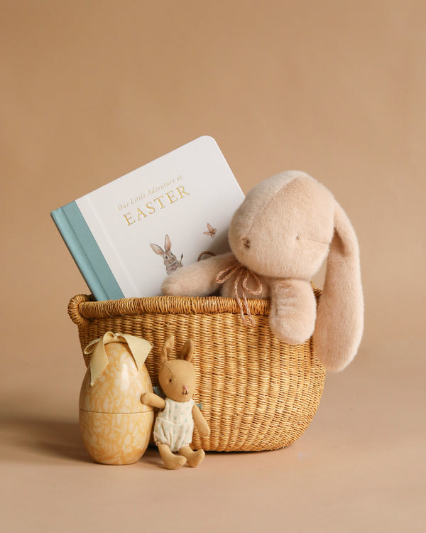 The Maileg Easter Basket Set includes a Maileg plush bunny, the "Our Little Adventure at Easter" board book, a decorative egg, and a small bunny figurine—all arranged on a beige background—making it an ideal Easter gift.