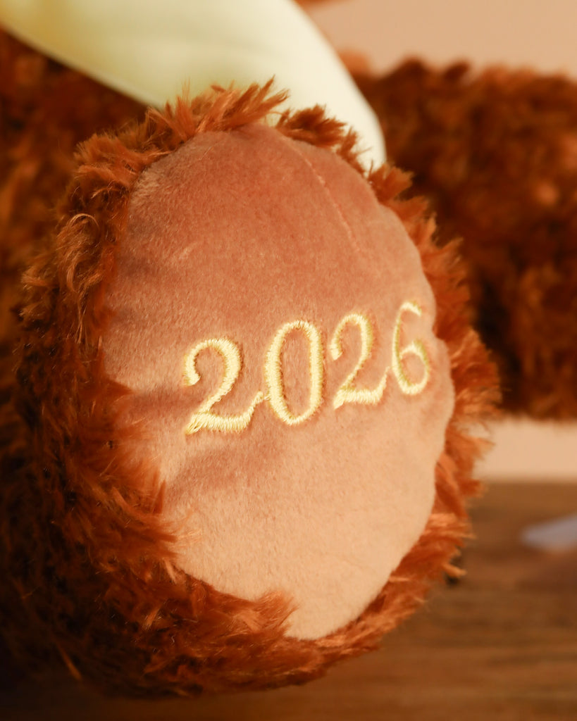 A close-up of the Steiff Cosy Year Bear 2026 13" shows its paw embroidered with “2026” in yellow thread on a wooden surface—a charming keepsake plush and thoughtful New Year gift.