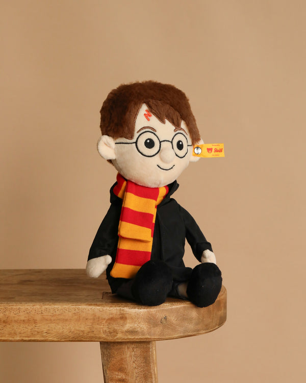 The Steiff Harry Potter Plush 11", featuring round glasses, a lightning bolt scar, and an official Gryffindor scarf, sits on a wooden bench against a plain beige background.