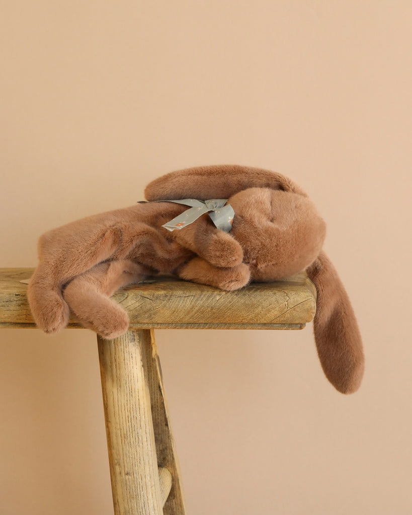 A Maileg Sleeping Bunny in Carry Cot with floppy ears and a silver ribbon rests on its side atop a rustic wooden stool against a beige background.