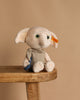 The Steiff Harry Potter “Dobby” with Sock 10" plush, featuring large ears, green eyes, a beige outfit, and an orange tag on its left ear, sits on a wooden stool against a plain beige background.