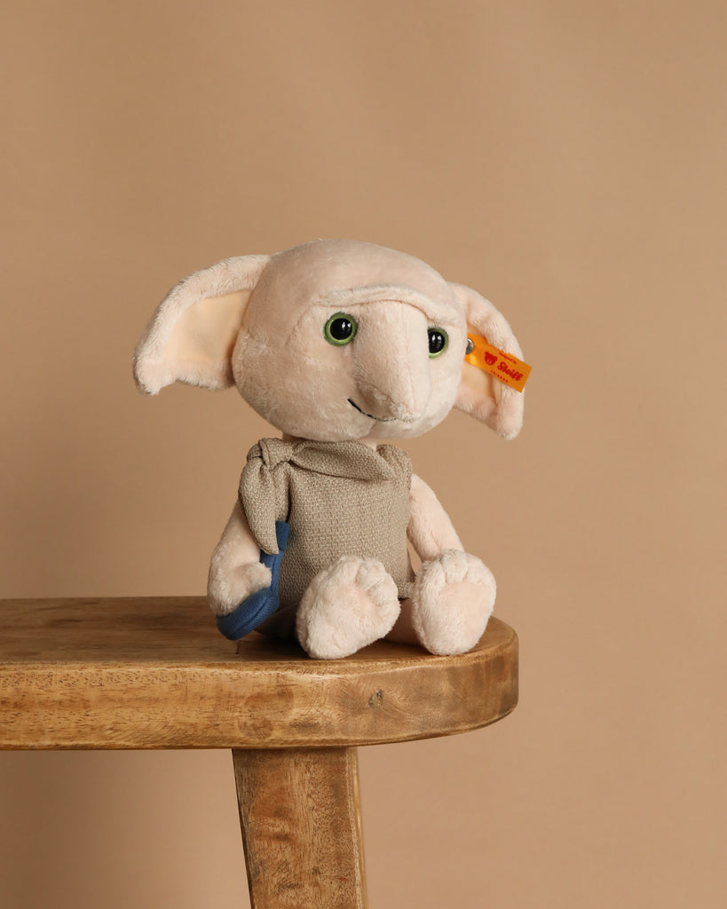 The Steiff Harry Potter “Dobby” with Sock 10" plush, featuring large ears, green eyes, a beige outfit, and an orange tag on its left ear, sits on a wooden stool against a plain beige background.
